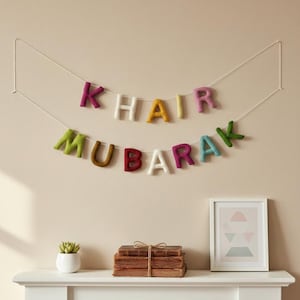 Op de afbeelding: Een viltbanner met de woorden "Khair Mubarak" in kleurrijke letters geregen aan een witte draad. De banner hangt aan een beige muur boven een witte schoorsteenmantel. Een kleine plant, boeken en een ingelijste prent staan op de schoorsteenmantel.