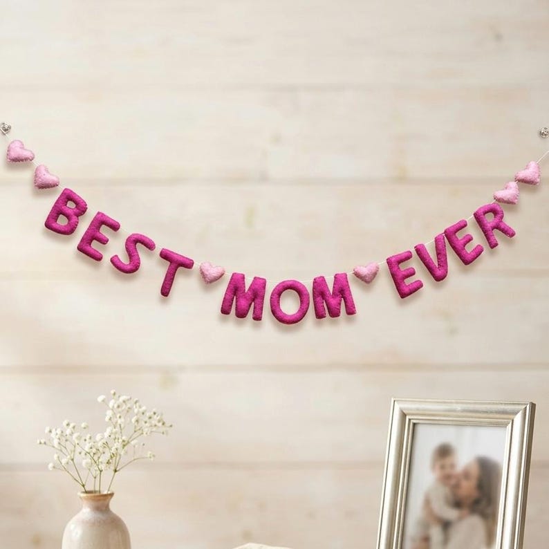 May include: A pink felt banner that reads "BEST MOM EVER" with heart-shaped accents. The banner is strung across a light-colored wooden wall. A framed photo and a vase of flowers are in the foreground.