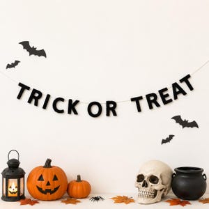 以下が含まれることがあります： 「TRICK OR TREAT」のバナー、カボチャ、骸骨、大釜が特徴のハロウィーンをテーマにしたディスプレイ。黒いコウモリが白い背景を飾っています。オレンジ色のジャック・オー・ランタンと小さなカボチャが前景にあり、黒いランタンがあります。