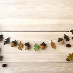 May include: A felt garland featuring acorns and leaves in shades of brown, tan, green, and black. The garland is strung on a white string and is set against a light wood background. Perfect for fall decor.