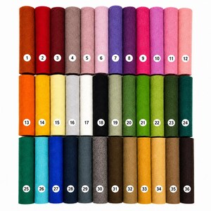 May include: A collection of thirty-six cylindrical rolls of felt fabric arranged in three rows of twelve. Each roll is a distinct solid color, ranging from warm reds and oranges to cool blues, greens, and neutrals, each labeled with a white numbered circle.