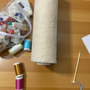 Peut inclure: Composition de fournitures de bricolage comprenant un rouleau de feutre beige, des bobines de fil de différentes couleurs et un sac transparent rempli de perles et de petits objets artisanaux. Un outil en bois et des ciseaux sont également visibles.