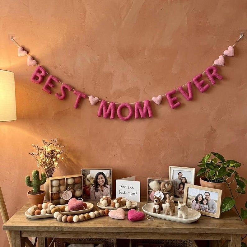 May include: A decorative display featuring a pink felt banner that reads "BEST MOM EVER" with heart accents. The display includes framed photos, felt ornaments, and a small potted cactus, all arranged on a wooden table.