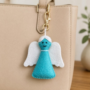 May include: A felt angel keyring featuring a turquoise triangular body and white wings. The angel has a white hair detail, two small black dot eyes, and a stitched black mouth. It is attached to a gold-toned metal lobster clasp hook.