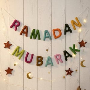 Op de afbeelding: Een feestelijke Ramadan Mubarak banner met kleurrijke viltletters, sterren en manen. De banner is geregen met lichtslingers tegen een witte houten achtergrond, wat een warme en feestelijke sfeer creëert.