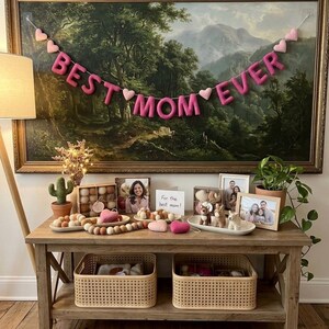 May include: A wooden table displays decorative items, including framed photos, felt hearts, and a banner that reads "BEST MOM EVER." A painting of a landscape hangs above the table. Two woven baskets sit below.