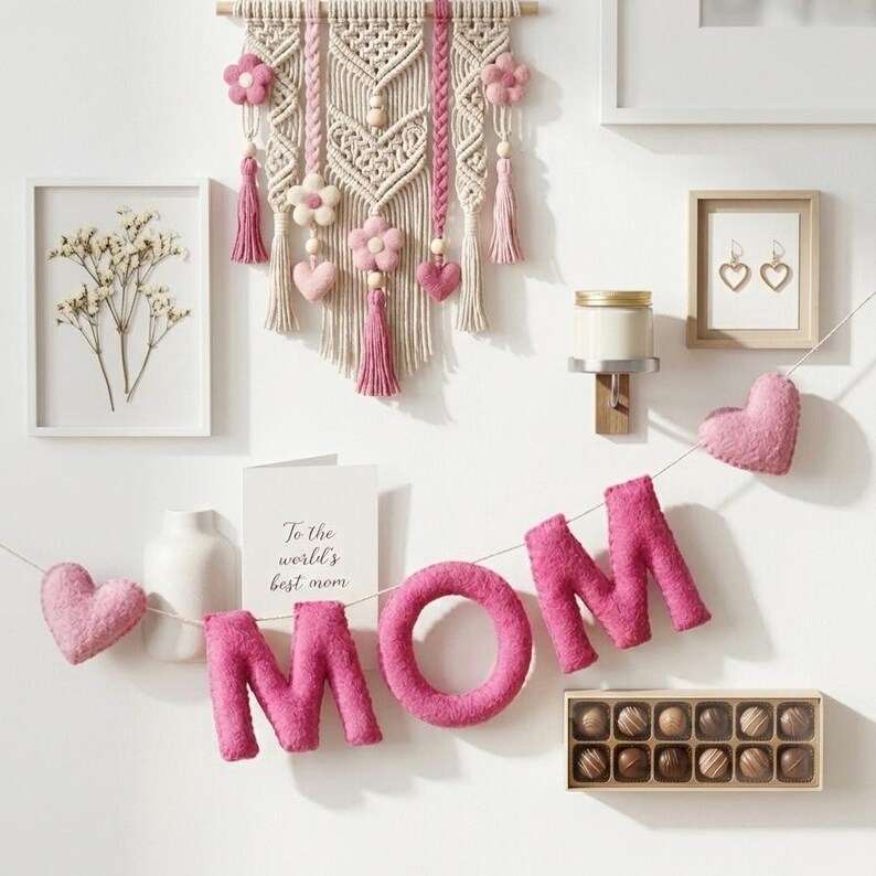May include: A wall display featuring a macrame hanging, framed art, and a felt garland spelling "MOM." The macrame includes pink flowers and tassels. A card reads "To the world's best mom." A box of chocolates is also present.