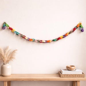 Puede incluir: Una colorida guirnalda de cadena de fieltro, suspendida contra una pared blanca. La guirnalda presenta una variedad de colores, incluyendo rojo, naranja, amarillo, verde, azul y morado. La guirnalda cuelga sobre una mesa de madera con un jarrón y objetos decorativos.