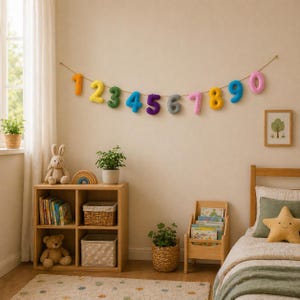 May include: A vibrant felt number garland, displaying the digits 1 to 0 in assorted colours, suspended on twine. The banner is hung on a neutral wall in a child's bedroom, above a wooden bookshelf and bed, creating a playful atmosphere.