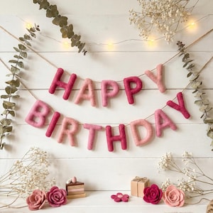 Può includere: Una ghirlanda di lettere in feltro che compone la scritta HAPPY BIRTHDAY in tonalità rosa e rosso, appesa a un filo sottile. Le lettere sono morbide e testurizzate. Sotto ci sono fiori in feltro, una torta in legno e un piccolo pacchetto.
