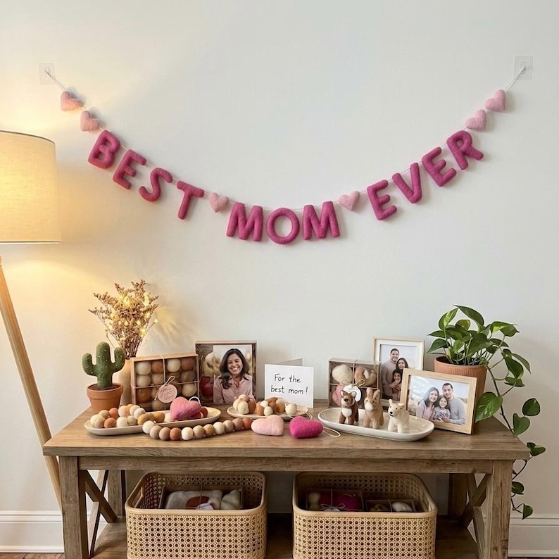May include: A wooden table displays a "BEST MOM EVER" banner, photos, and decorative items. The banner is made of pink felt letters and hearts. Other items include framed photos, small figurines, and a potted plant, creating a celebratory atmosphere.