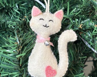 May include: A white felt cat Christmas ornament with pink ears and a pink heart. The cat has a pink ribbon collar with a silver bell. The ornament is hanging on a green Christmas tree.