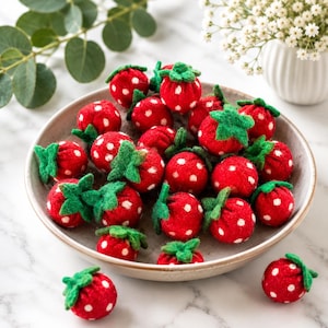 Op de afbeelding: Een verzameling rode vilt aardbeien met witte stippen en groene bladeren, gerangschikt in een grijze keramische kom. De aardbeien zijn een ambachtelijk item, mogelijk voor decoratie.
