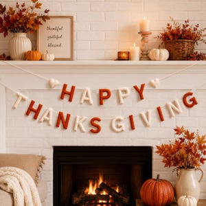以下が含まれることがあります： オレンジとクリーム色の文字が交互に並ぶHAPPY THANKSGIVINGのフェルトガーランド。小さなハートのアクセントが付いた白い紐で吊るされています。このバナーは、カボチャで飾られた白いレンガの暖炉の棚の前にディスプレイされています。