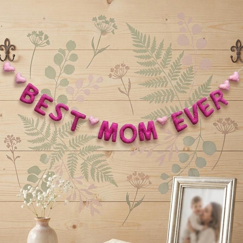 May include: A felt banner with the words "BEST MOM EVER" in magenta letters, strung with pink heart-shaped accents. The banner hangs against a light wood background with floral stencils and decorative hooks.