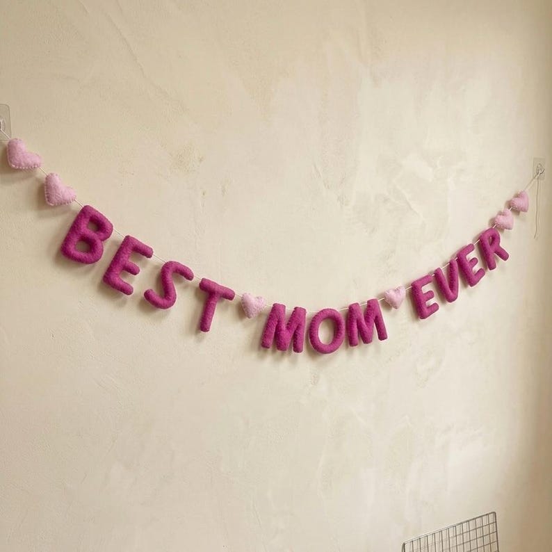 May include: A felt banner with the words "BEST MOM EVER" in magenta letters, strung with small pink hearts. The banner is hanging against a light-colored wall, creating a festive and celebratory display.