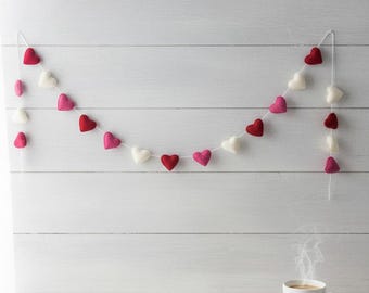 May include: A felt heart garland in shades of red, pink, and white hangs against a white wooden background. The garland is strung on a white string and is a decorative item. A white mug of coffee sits below the garland.