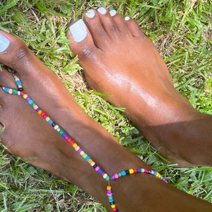 May include: A pair of feet with white nail polish on the toes. The feet are wearing a colorful beaded anklet that wraps around the ankle and between the toes.