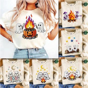 以下が含まれることがあります： 様々な刺繍デザインが施された、クリーム色のハロウィーンをテーマにしたスウェットシャツ。カボチャの焚き火の周りにいる幽霊、星と幽霊、焚き火や「BOO！」のサインを持っている幽霊などが描かれています。
