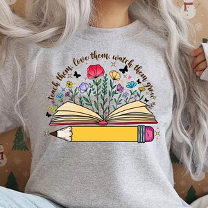 K&ouml;nnte beinhalten: Ein graues Sweatshirt mit einem grafischen Design eines Buches mit Blumen und einem Bleistift. Der Text auf dem Shirt lautet "Teach them, love them, watch them grow."