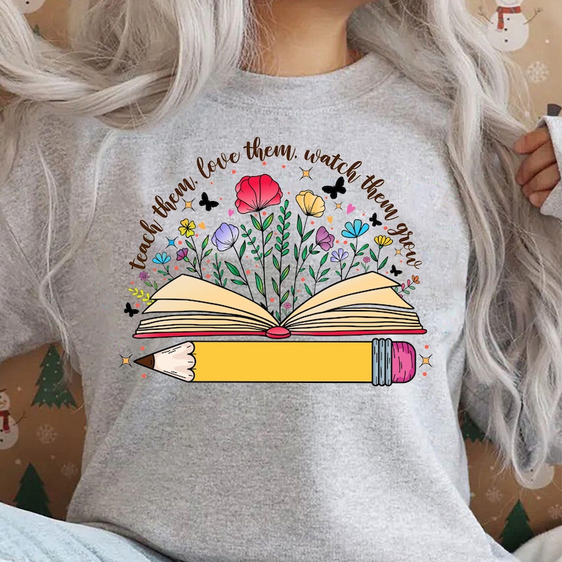K&ouml;nnte beinhalten: Ein graues Sweatshirt mit einem grafischen Design eines Buches mit Blumen und einem Bleistift. Der Text auf dem Shirt lautet "Teach them, love them, watch them grow."