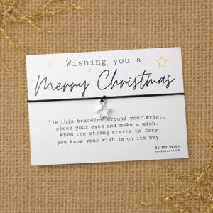 May include: A black string bracelet with a silver star charm attached to a white card. The card features the text Wishing you a Merry Christmas and instructions to tie the bracelet, close your eyes, and make a wish. It includes the text BE MY WISH handmade in UK.