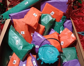 May include: A Christmas advent calendar with 24 individually wrapped gifts. The gifts are wrapped in orange, purple, and green tissue paper. The gifts are numbered 1 through 24. The calendar is decorated with a black cauldron with a teal lid and a red and green Christmas ornament.