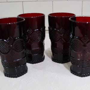 May include: A set of four deep red glass tumblers featuring a raised diamond and circular geometric pattern. Each cylindrical drinking glass has a textured base and a smooth rim, showcasing a consistent dark ruby color throughout the glass body.