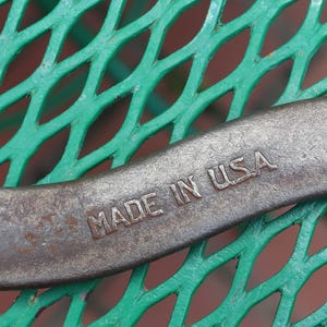 May include: A vintage, curved wrench made of metal, with the text "MADE IN USA" stamped on the body. The wrench is resting on a green, diamond-patterned surface. The numbers "838" and "16" are also visible.