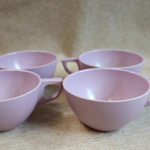 May include: Four light pink cups with handles are arranged in a group. The cups have a simple, rounded design and a matte finish. The handles are rectangular and attached to the side of the cups.
