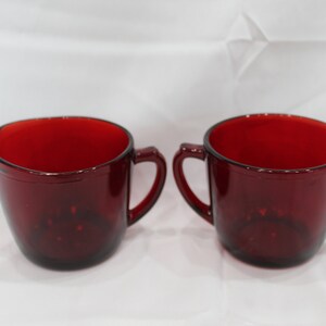 May include: Two ruby red glass cups with handles. One cup has a spout. The cups are made of a transparent glass material. The cups are likely part of a vintage glassware set.