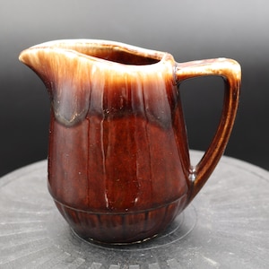 Pode incluir: Um pequeno creamer de cerâmica marrom e branco com alça. O creamer tem um acabamento esmaltado e uma forma ligeiramente arredondada.
