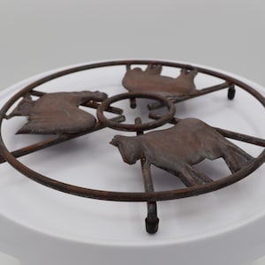 May include: A rustic metal trivet with a circular design featuring three stylized cows. The trivet is made of dark brown metal and has a distressed finish.