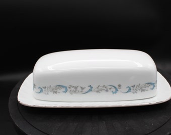 Könnte beinhalten: Eine weiße Porzellan-Butterdose mit einem blauen und grauen Blumendesign. Die Dose hat einen Deckel und einen Sockel.