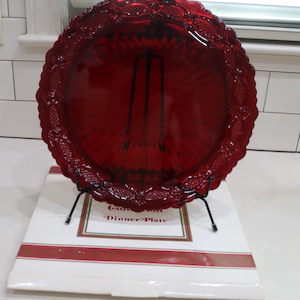 Könnte beinhalten: Ein runder, rubinroter Glasteller mit einem dekorativen, gewellten Rand und einem strukturierten Gittermuster. Der Teller steht auf einem schwarzen Metallständer vor einer weißen Schachtel mit der Aufschrift "Dinner Plate".