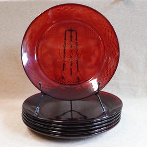 May include: Set of six ruby red glass plates. Each plate features a diamond pattern around the rim and a decorative design in the center. The plates are displayed on a black metal stand, showcasing their rich color and design.