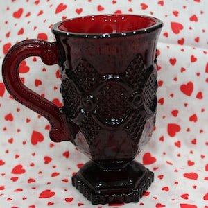 May include: A dark red glass mug with a footed base and a detailed design. The mug has a handle and is made of ruby red glass.