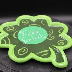 May include: Green shamrock-shaped ceramic dish with a green circle in the center that says "Kiss for luck" in white lettering. The dish has a raised rim and a glossy finish.