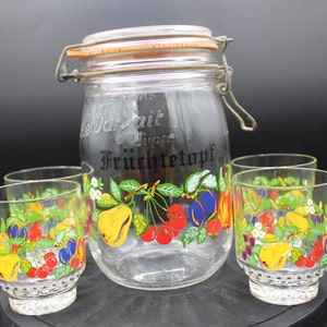 May include: A clear glass jar with a lid and a black and white label that says "Fruchtettopf" with a design of fruit around the jar. There are four clear glass cups with a fruit design around them.