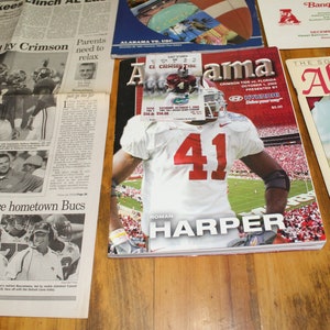 Pode incluir: Uma coleção de memorabilia esportiva, incluindo jornais com manchetes como "Crushed by Crimson" e revistas com jogadores de futebol. Um bilhete e um convite para um banquete também são visíveis.