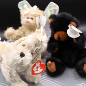 Könnte beinhalten: Drei Plüschtiere: ein weißer Hund mit einer rosa Schleife, ein brauner Bär mit schwarzen Flügeln und ein schwarzer Bär mit braunen Ohren und einer braunen Nase.