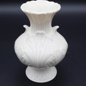 May include: A white ceramic vase with an ornate design. The vase has a wide base and a narrow neck.