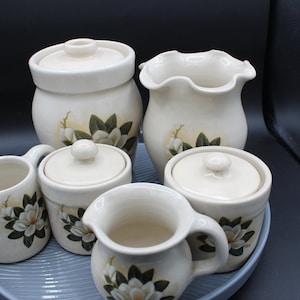 Könnte beinhalten: Ein Set aus sechs weißen Keramik-Küchenbehältern mit Blumendesigns. Die Behälter sind auf einem blauen Tablett angeordnet. Die Behälter haben Deckel und sind mit weißen Blumen und grünen Blättern verziert.