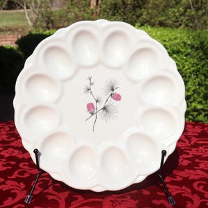 May include: A white ceramic egg plate with a scalloped edge and 12 egg indentations. The centre features a black and pink floral design. The plate is displayed on a black metal stand, ideal for serving eggs.