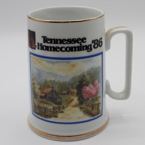 May include: White ceramic mug with a gold rim and handle. The mug features a colorful illustration of a rural scene with a house, trees, and a path. The text "Tennessee Homecoming '86" is printed in black on the mug.