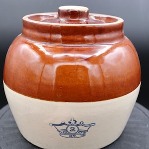 Op de afbeelding: Een bruine en witte geglazuurde keramische pot met een deksel. De pot heeft een blauw en wit kroonontwerp met het nummer "2" en de tekst "QT." aan de voorkant.