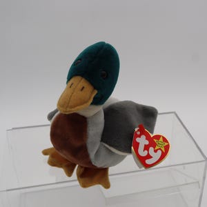 May include: A plush duck toy with a green head, yellow beak, brown body, gray wings, and yellow feet. It has a red heart-shaped tag with white text and a yellow star.