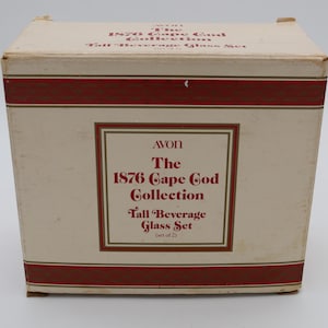 May include: A rectangular box with the text "AVON The 1876 Cape Cod Collection Tall Beverage Glass Set (set of 2)". The box is off-white with a dark red band around the top and bottom.