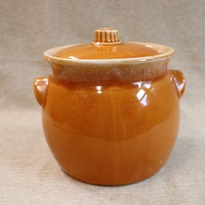 May include: A glazed ceramic pot with a lid. The pot is a warm, glossy orange-brown color with a lighter rim and a small handle on the lid. It has two small handles on the sides.