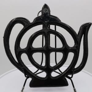 May include: Black cast iron teapot-shaped hot pad with a decorative design. The hot pad is designed to look like a teapot with a handle and spout.